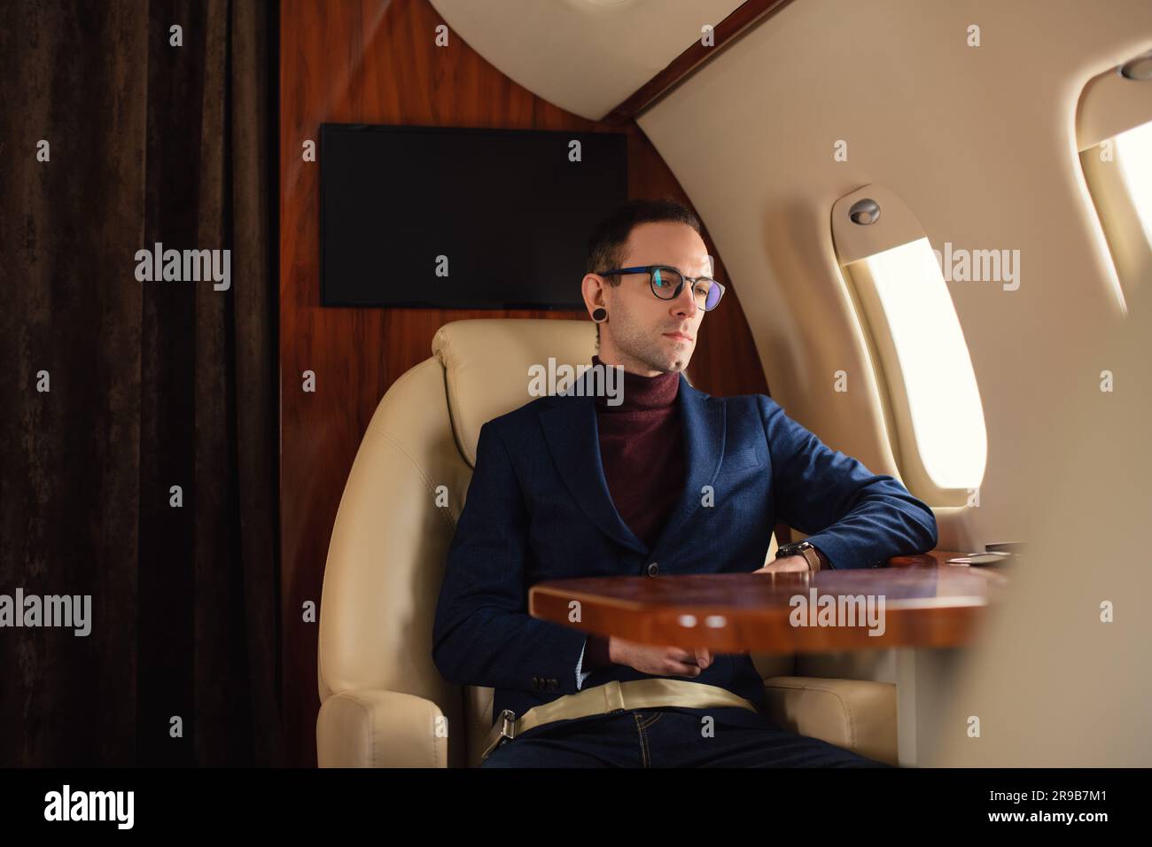 Junger, eleganter, selbstbewusster männlicher Geschäftsmann mit Brille in blauer Jacke, der durch ein Firmenflugzeugfenster blickt, Geschäftskonzept für Reisen und Fluggesellschaften Stockfoto