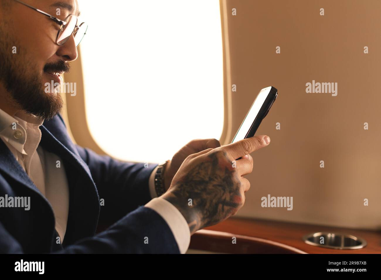 Eleganter junger asiatischer Geschäftsführer, Geschäftsmann mit Brille und Anzug, der im ersten Klasse eines Privatjet sitzt und mit einem modernen Smartphone arbeitet Stockfoto