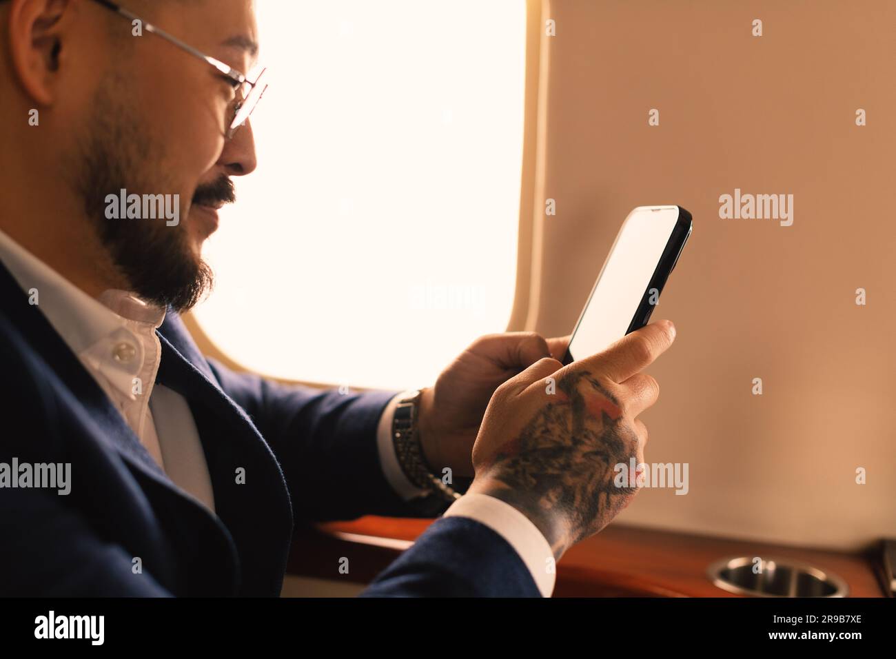Eleganter junger asiatischer Geschäftsführer, Geschäftsmann mit Brille und Anzug, der im ersten Klasse eines Privatjet sitzt und mit einem modernen Smartphone arbeitet Stockfoto