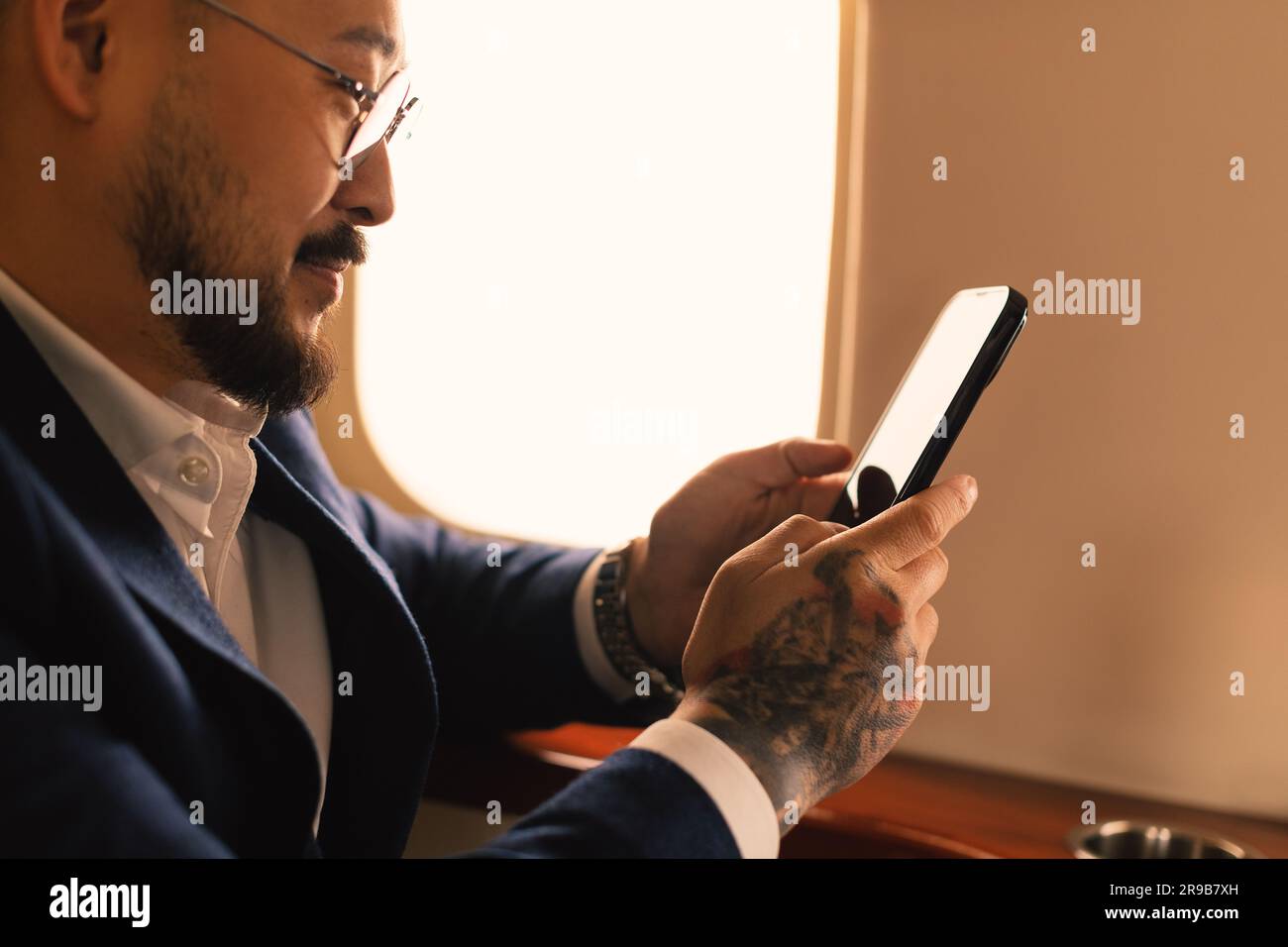 Eleganter junger asiatischer Geschäftsführer, Geschäftsmann mit Brille und Anzug, der im ersten Klasse eines Privatjet sitzt und mit einem modernen Smartphone arbeitet Stockfoto