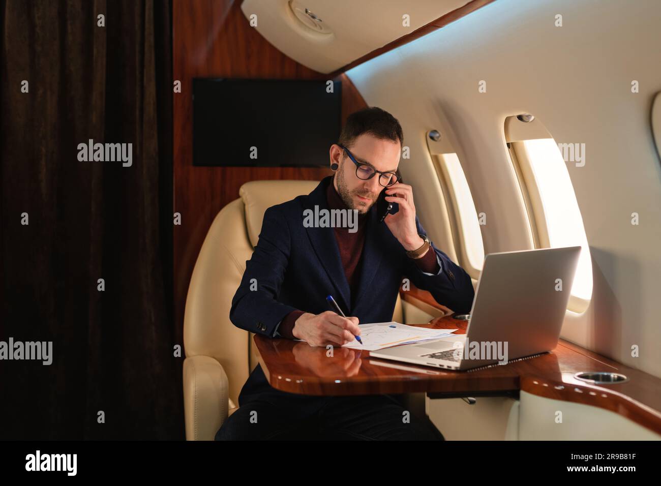 Junger eleganter Geschäftsmann mit Brille, der in einem Privatflugzeug sitzt und in Papierdokumenten arbeitet, die mit dem Handy und Laptop sprechen Stockfoto