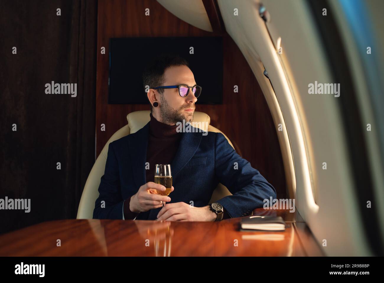Eleganter junger Geschäftsführer mit Brille in Jacke sitzt in einem privaten Flugzeug Jet und trinkt Champagner während des Fluges Stockfoto