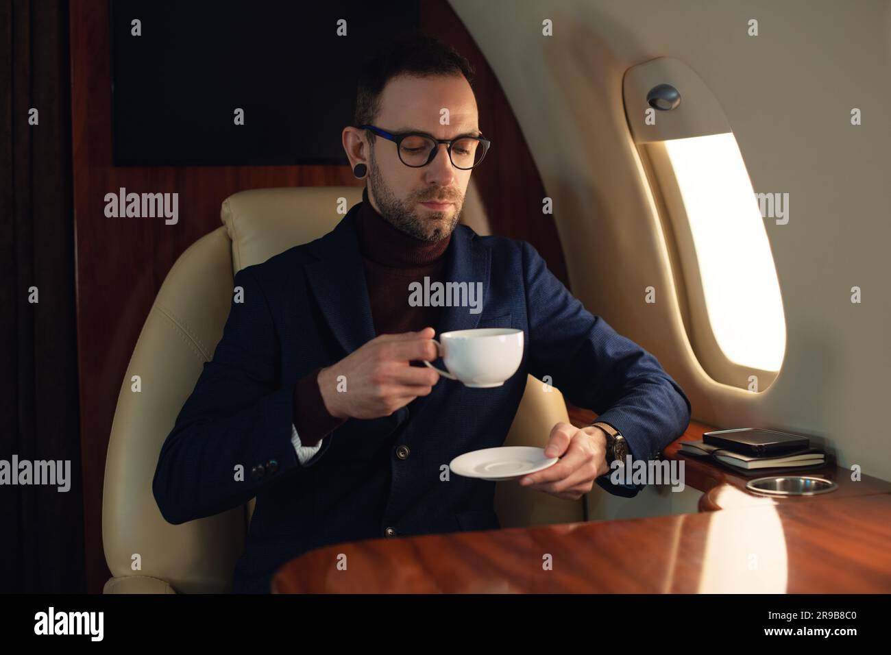 Eleganter CEO junger Geschäftsmann mit Brille eine Jacke, die in einem erstklassigen Privatjet sitzt, Kaffee genießt und aus dem Fenster schaut Stockfoto