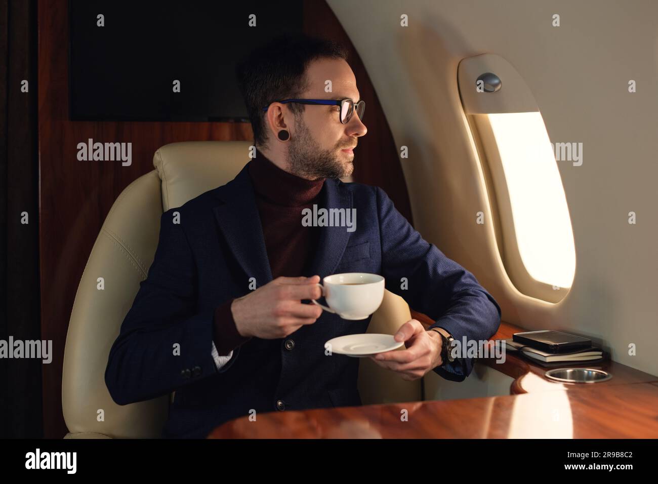 Eleganter CEO junger Geschäftsmann mit Brille und Jacke, der im ersten Klasse eines Privatjets sitzt und Kaffee genießt und aus dem Fenster blickt Stockfoto