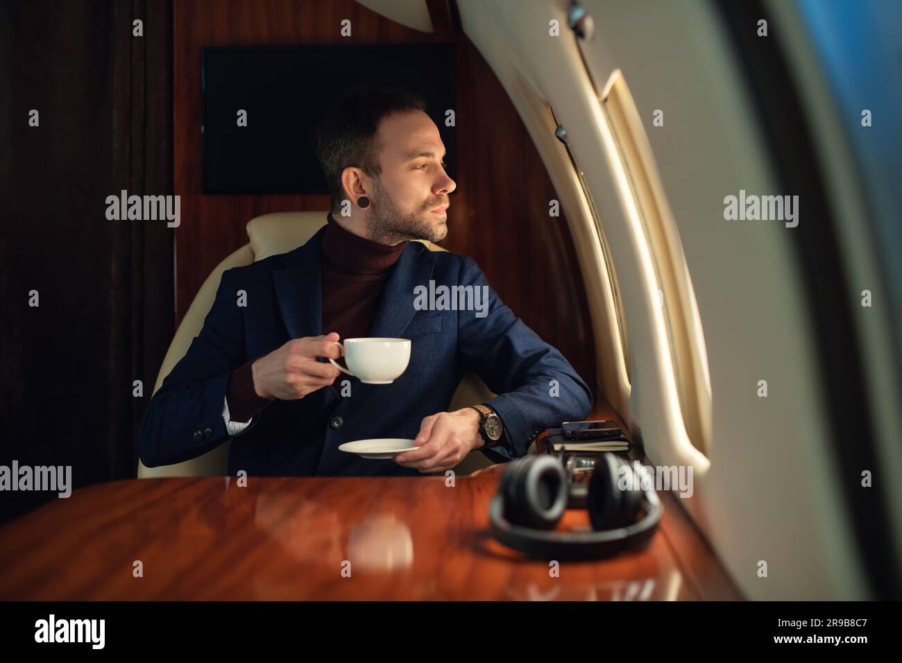 Eleganter CEO, junger Geschäftsmann in einer Jacke, sitzt in einem erstklassigen Privatjet und genießt Kaffee und schaut aus dem Fenster Stockfoto