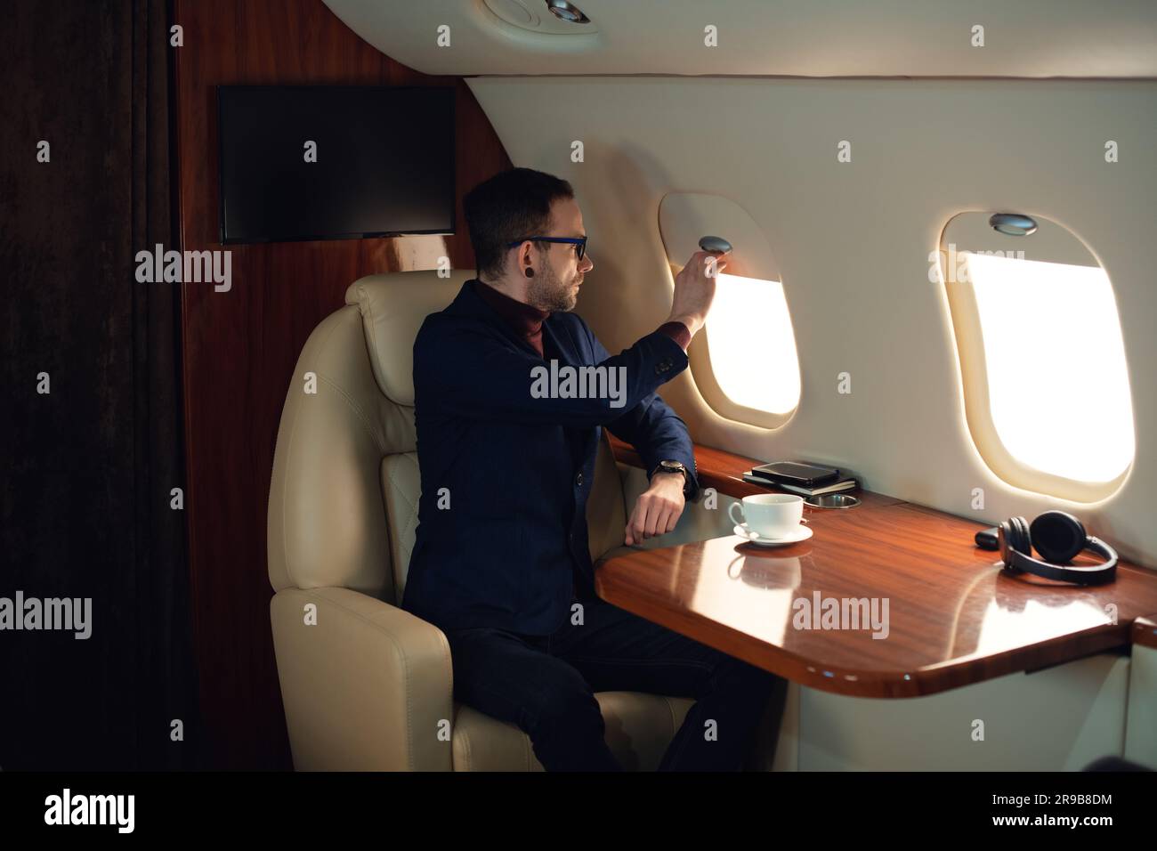 Der elegante Geschäftsmann für Erwachsene mit Brille zieht vor dem Start in einem privaten Businessjet den Fensterschirm herunter, der selbstbewusste Geschäftsmann in einer blauen Jacke Stockfoto