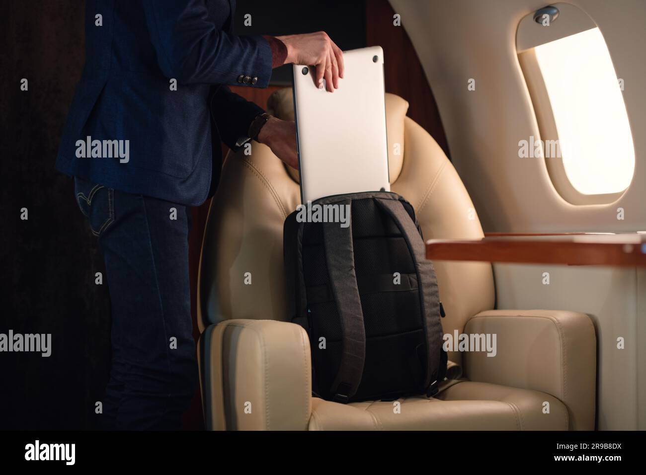 Ein eleganter Geschäftsmann aus der Nähe zieht einen modernen Laptop heraus und stellt ihn auf den Tisch, um von dort aus in einem erstklassigen Privatflugzeug zu arbeiten Stockfoto