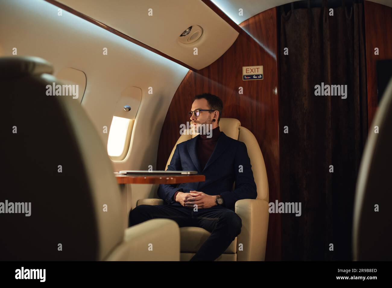 Junger, eleganter, selbstbewusster CEO-Geschäftsmann mit Brille in einer blauen Jacke, die durch ein erstklassiges Fenster im Flugzeug schaut Stockfoto