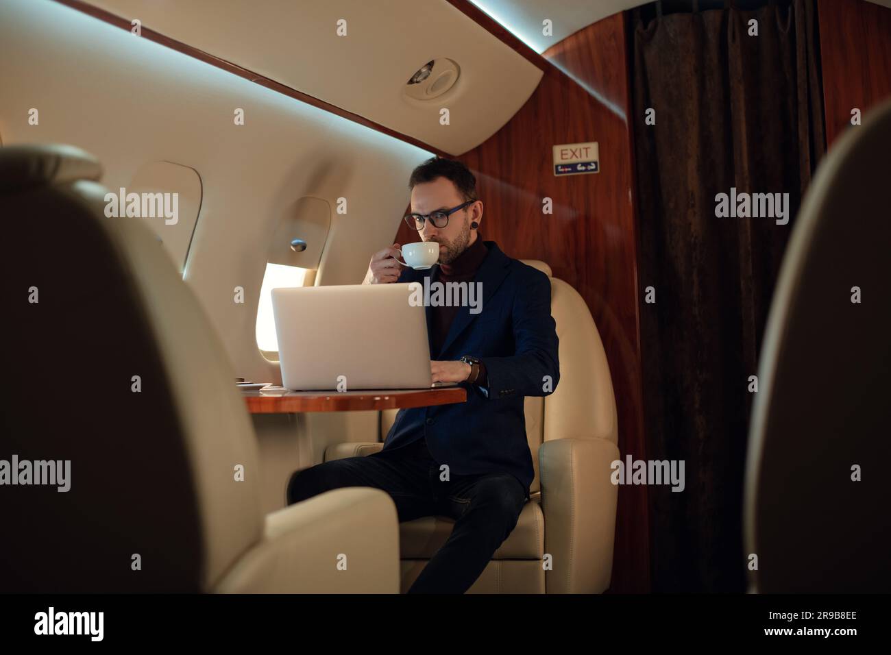 Eleganter CEO junger Geschäftsmann mit Brille Mann sitzt drinnen in erstklassigem Privatjet trinkt Kaffee, nachdem er an seinem modernen Laptop gearbeitet hat Stockfoto