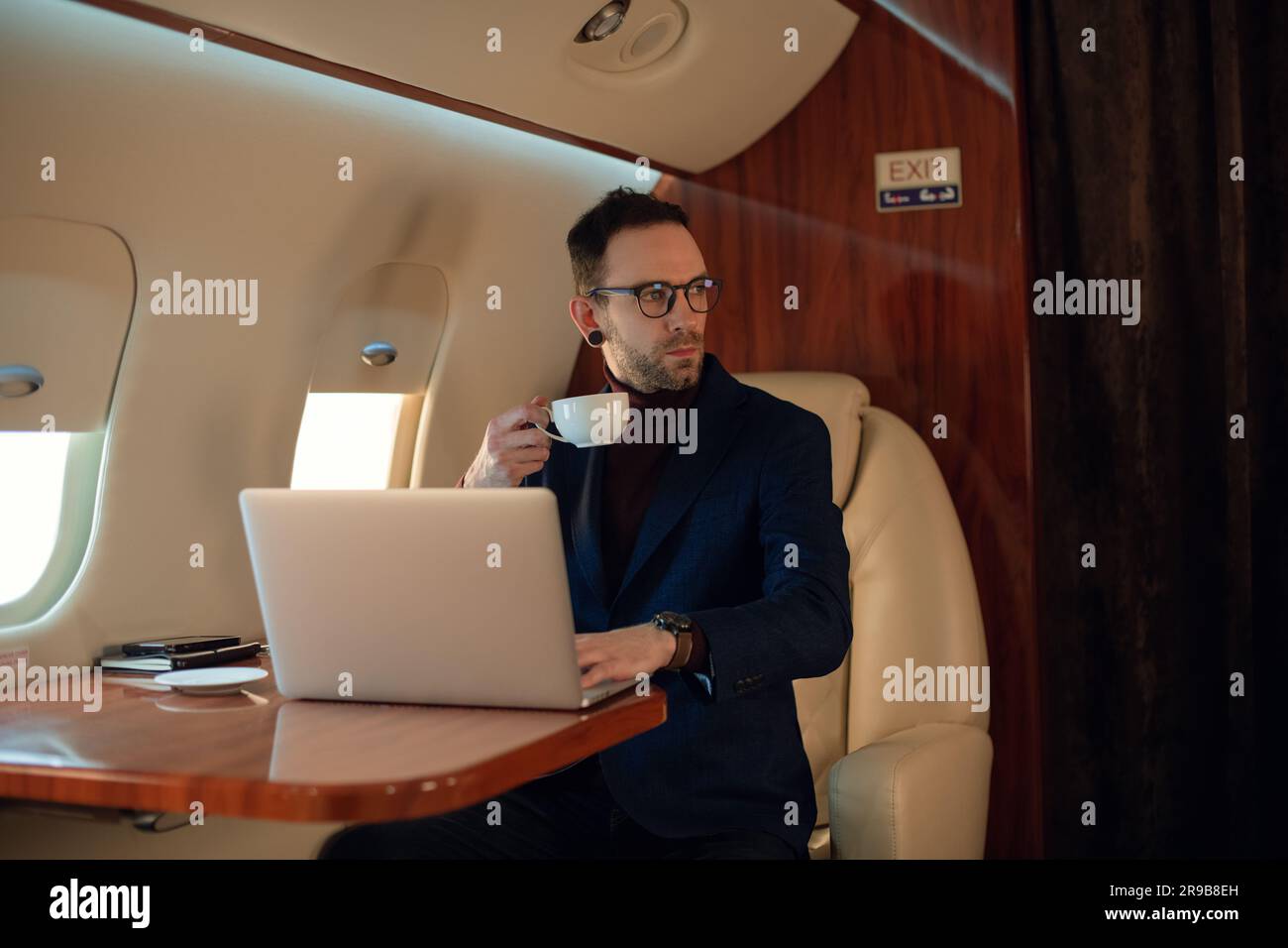 Eleganter CEO junger Geschäftsmann mit Brille Mann sitzt drinnen in erstklassigem Privatjet trinkt Kaffee, nachdem er an seinem modernen Laptop gearbeitet hat Stockfoto