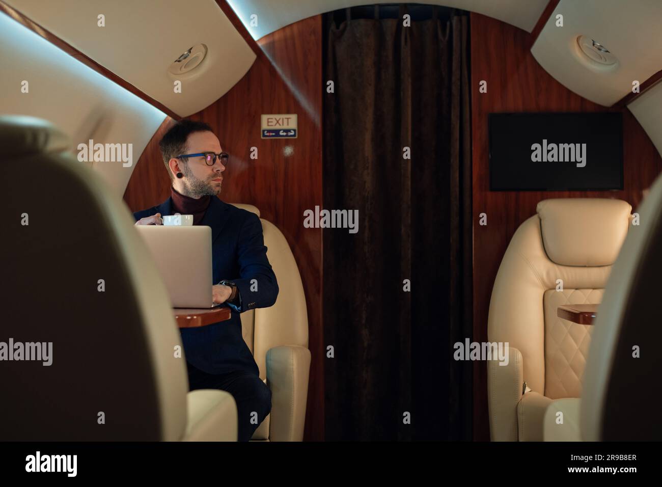 Eleganter CEO junger Geschäftsmann mit Brille Mann sitzt drinnen in erstklassigem Privatjet trinkt Kaffee, nachdem er an seinem modernen Laptop gearbeitet hat Stockfoto
