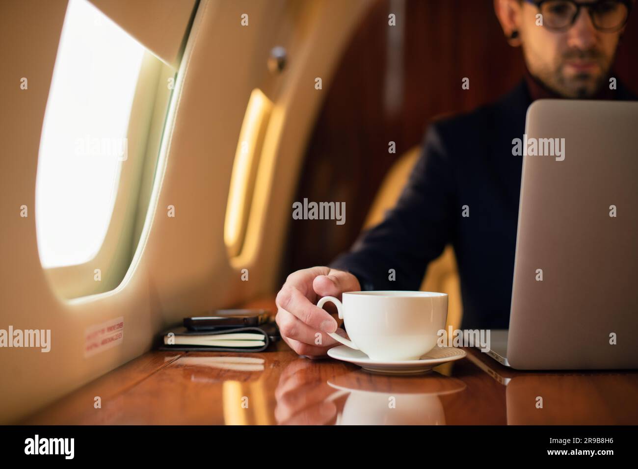 Nahaufnahme des Geschäftsmannes, der im privaten Flugzeug Jet nach einer Tasse Kaffee streckt, eleganter junger Geschäftsmann mit Brille, der im Privatjet sitzt Stockfoto
