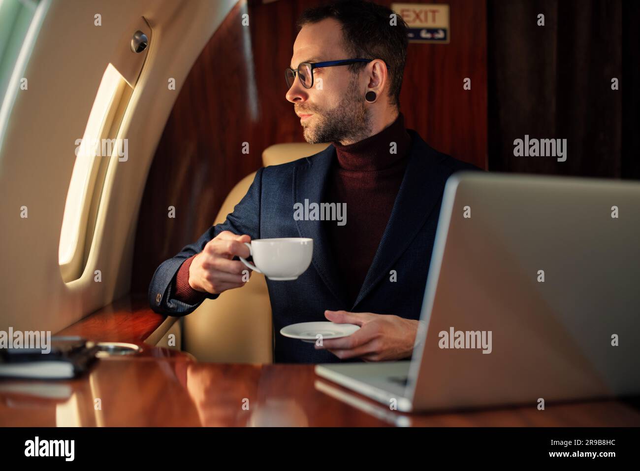 Eleganter CEO junger Geschäftsmann mit Brille Mann sitzt drinnen in erstklassigem Privatjet trinkt Kaffee, nachdem er an seinem modernen Laptop gearbeitet hat Stockfoto