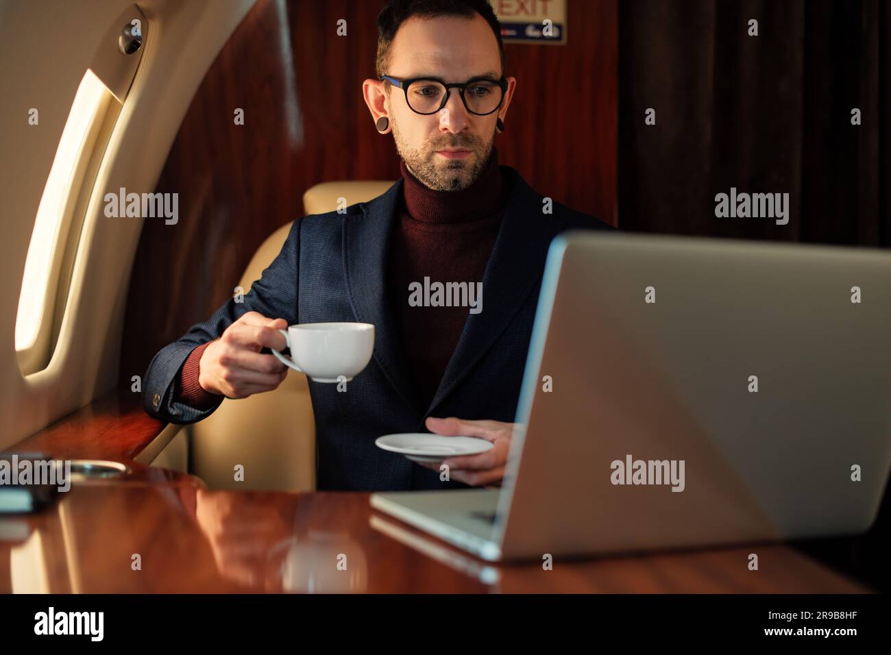Eleganter CEO junger Geschäftsmann mit Brille Mann sitzt drinnen in erstklassigem Privatjet trinkt Kaffee, nachdem er an seinem modernen Laptop gearbeitet hat Stockfoto