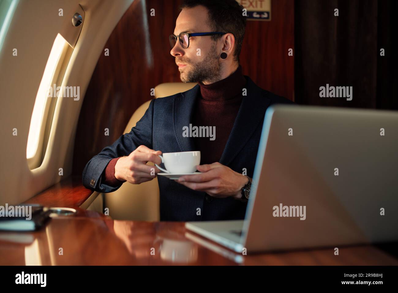 Eleganter CEO junger Geschäftsmann mit Brille Mann sitzt drinnen in erstklassigem Privatjet trinkt Kaffee, nachdem er an seinem modernen Laptop gearbeitet hat Stockfoto