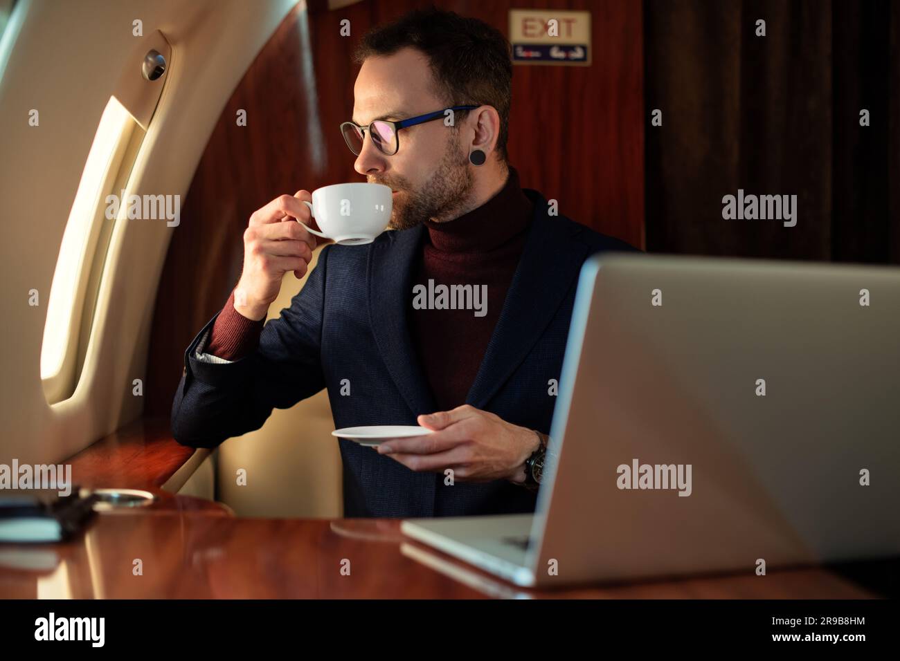 Eleganter CEO junger Geschäftsmann mit Brille Mann sitzt drinnen in erstklassigem Privatjet trinkt Kaffee, nachdem er an seinem modernen Laptop gearbeitet hat Stockfoto