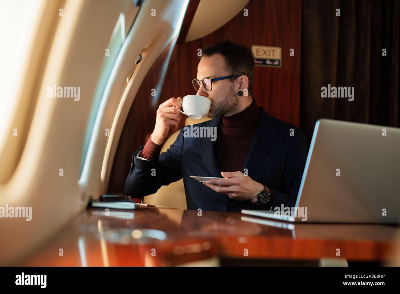 Eleganter CEO junger Geschäftsmann mit Brille Mann sitzt drinnen in erstklassigem Privatjet trinkt Kaffee, nachdem er an seinem modernen Laptop gearbeitet hat Stockfoto