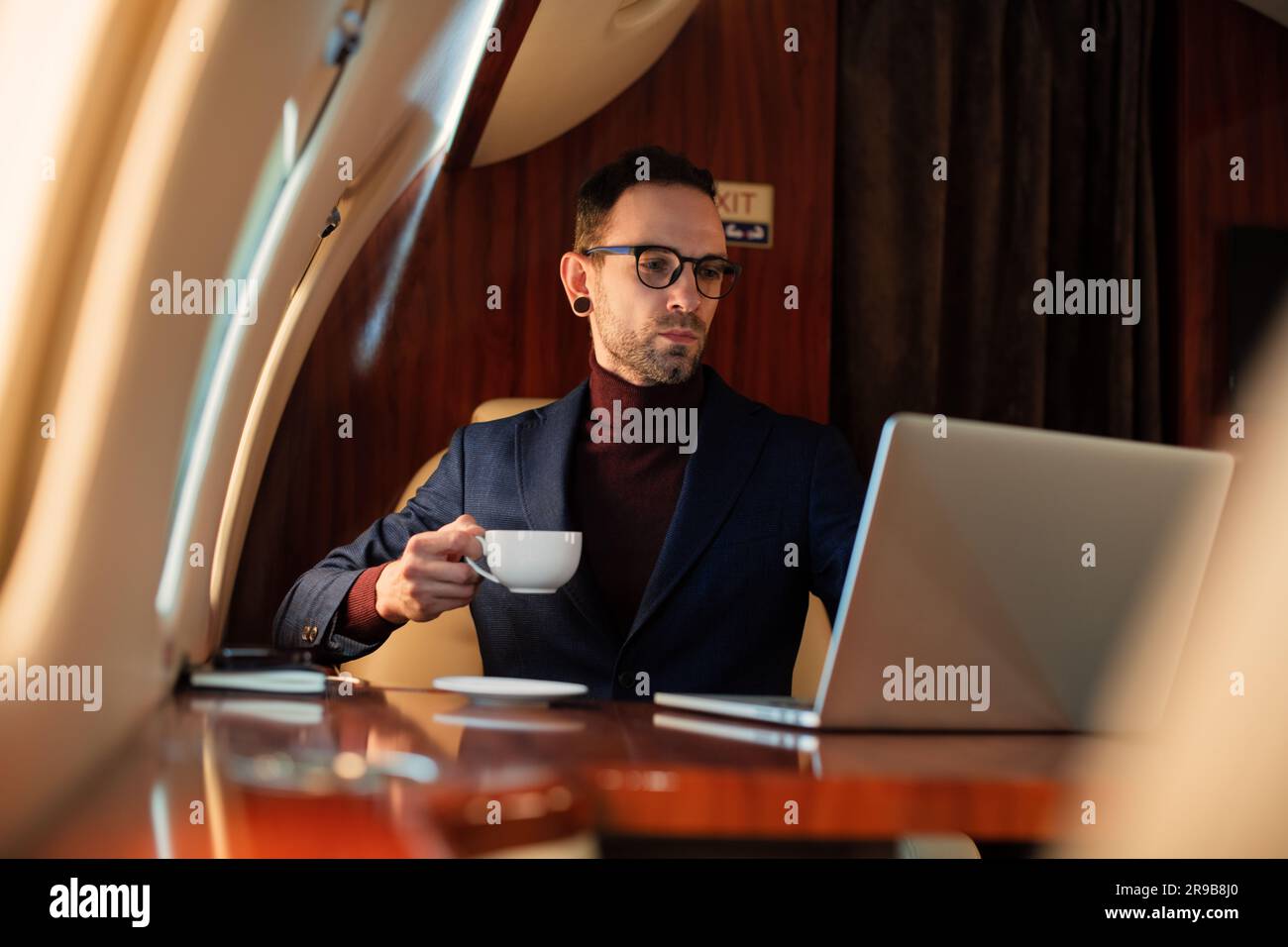 Eleganter CEO junger Geschäftsmann mit Brille Mann sitzt drinnen in erstklassigem Privatjet trinkt Kaffee, nachdem er an seinem modernen Laptop gearbeitet hat Stockfoto
