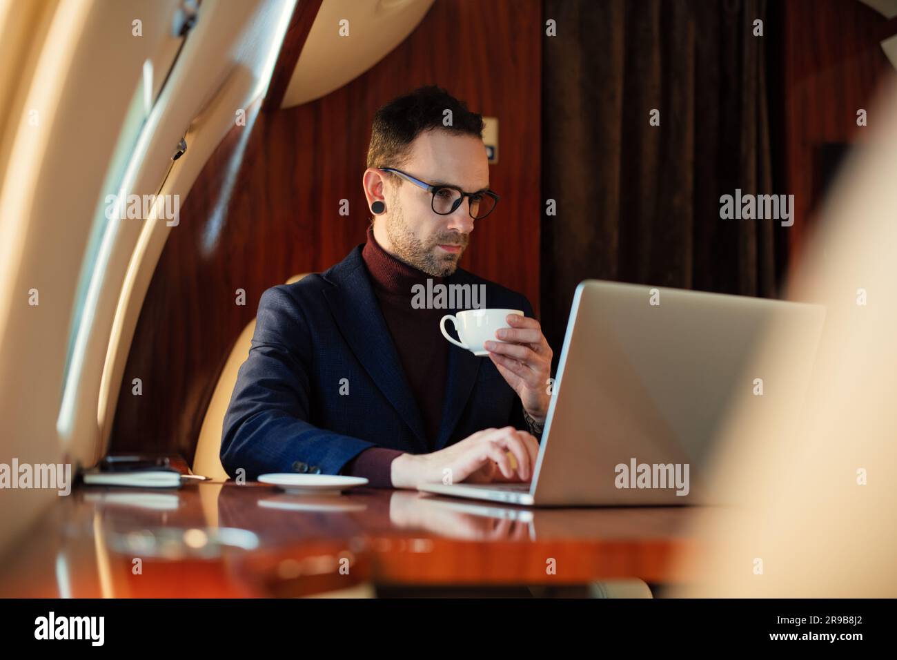 Eleganter CEO junger Geschäftsmann mit Brille Mann sitzt drinnen in erstklassigem Privatjet trinkt Kaffee, nachdem er an seinem modernen Laptop gearbeitet hat Stockfoto