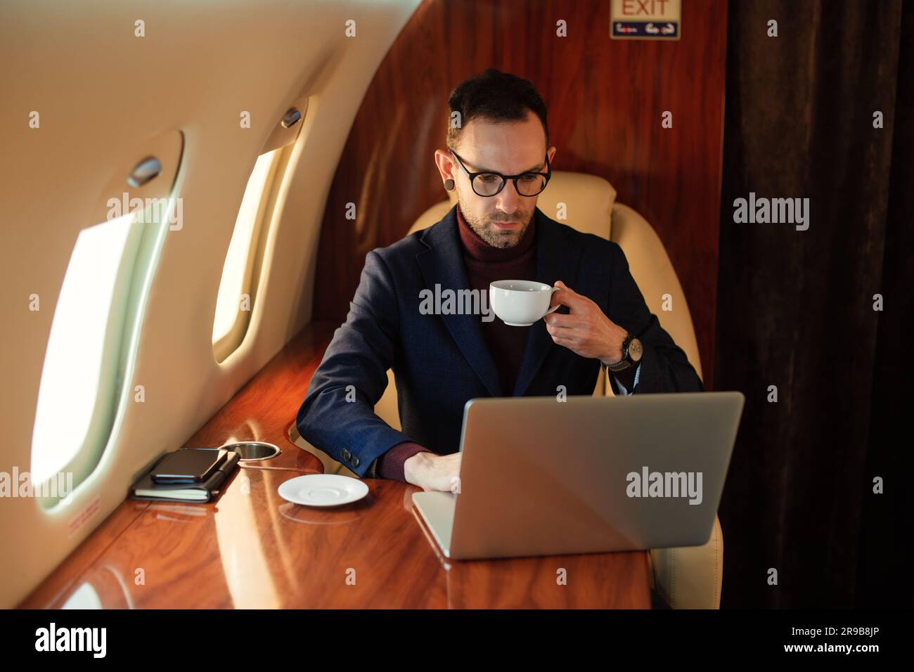 Eleganter CEO junger Geschäftsmann mit Brille Mann sitzt drinnen in erstklassigem Privatjet trinkt Kaffee, nachdem er an seinem modernen Laptop gearbeitet hat Stockfoto
