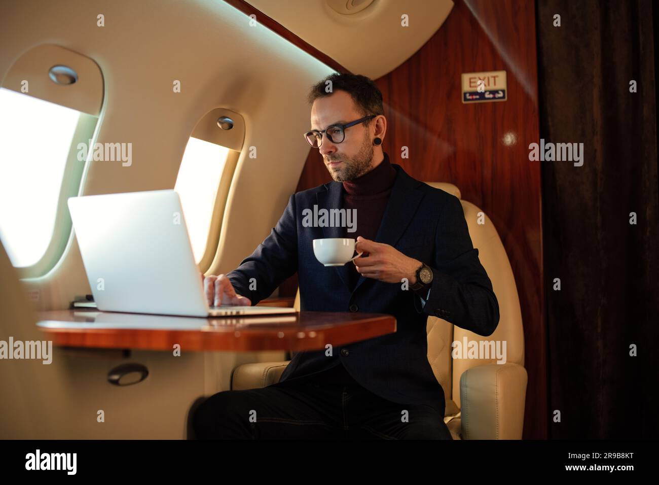 Eleganter CEO junger Geschäftsmann mit Brille Mann sitzt drinnen in erstklassigem Privatjet trinkt Kaffee, nachdem er an seinem modernen Laptop gearbeitet hat Stockfoto
