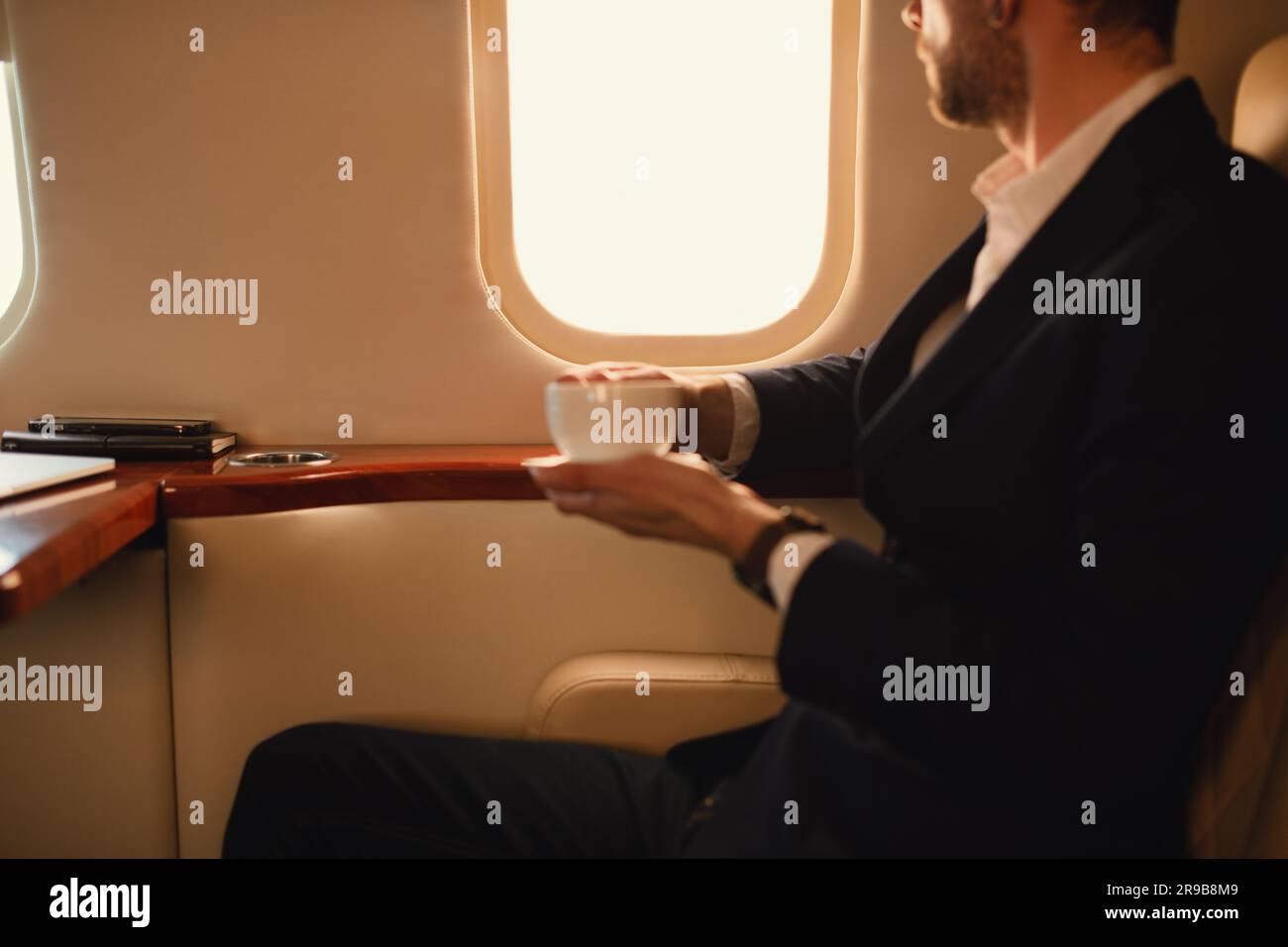 Ein nicht wiedererkennbarer, eleganter Geschäftsführer mit Brille in blauer Jacke, der in einem Privatflugzeug sitzt und Kaffee trinkt und durch ein Fenster schaut Stockfoto