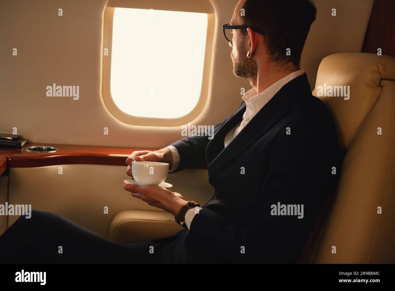 Ein nicht wiedererkennbarer, eleganter Geschäftsführer mit Brille in blauer Jacke, der in einem Privatflugzeug sitzt und Kaffee trinkt und durch ein Fenster schaut Stockfoto