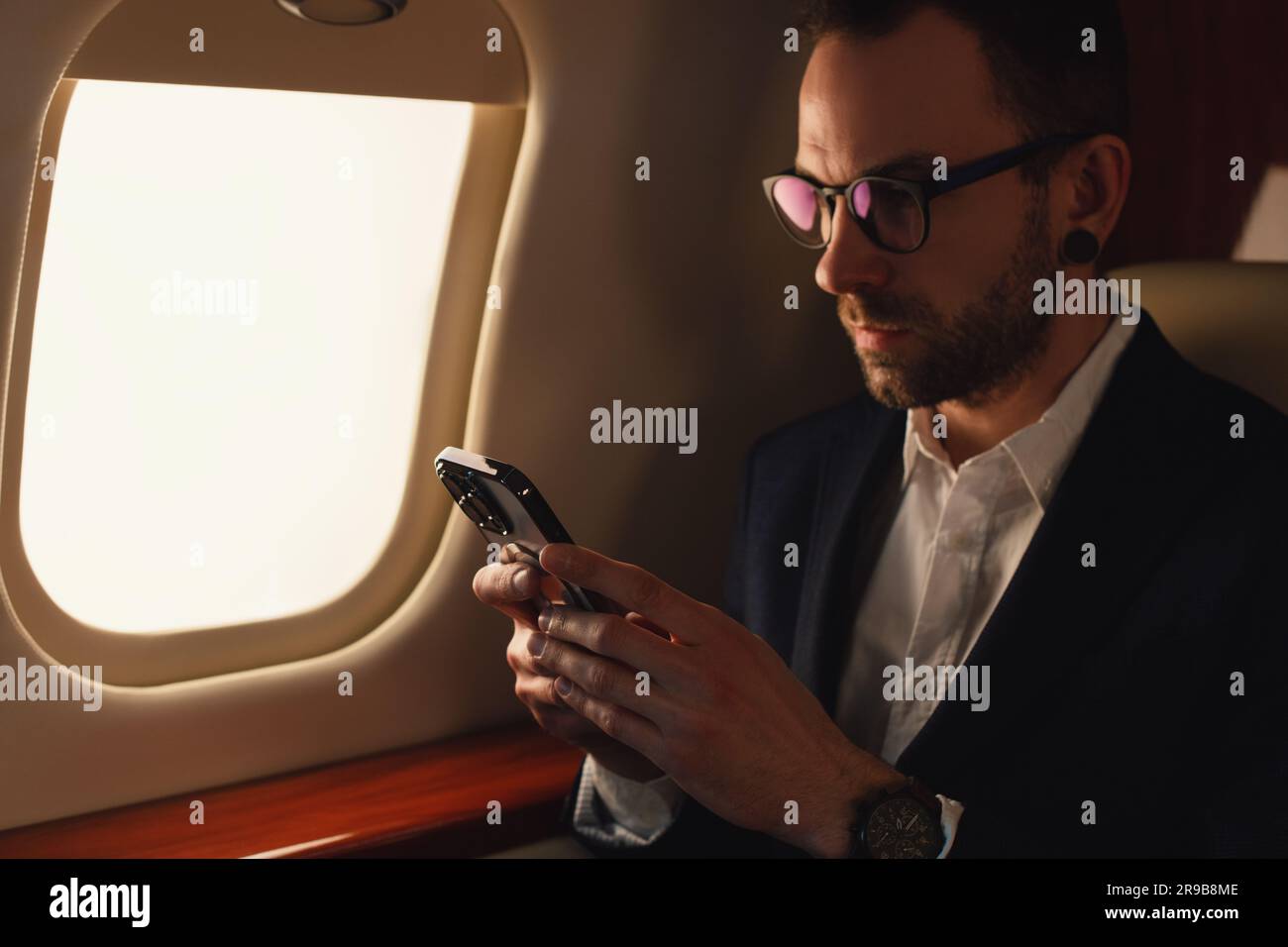 Eleganter CEO junger, selbstbewusster Geschäftsmann mit Brille Mann sitzt in einem erstklassigen Privatjet und arbeitet mit einem modernen Smartphone aus nächster Nähe Stockfoto