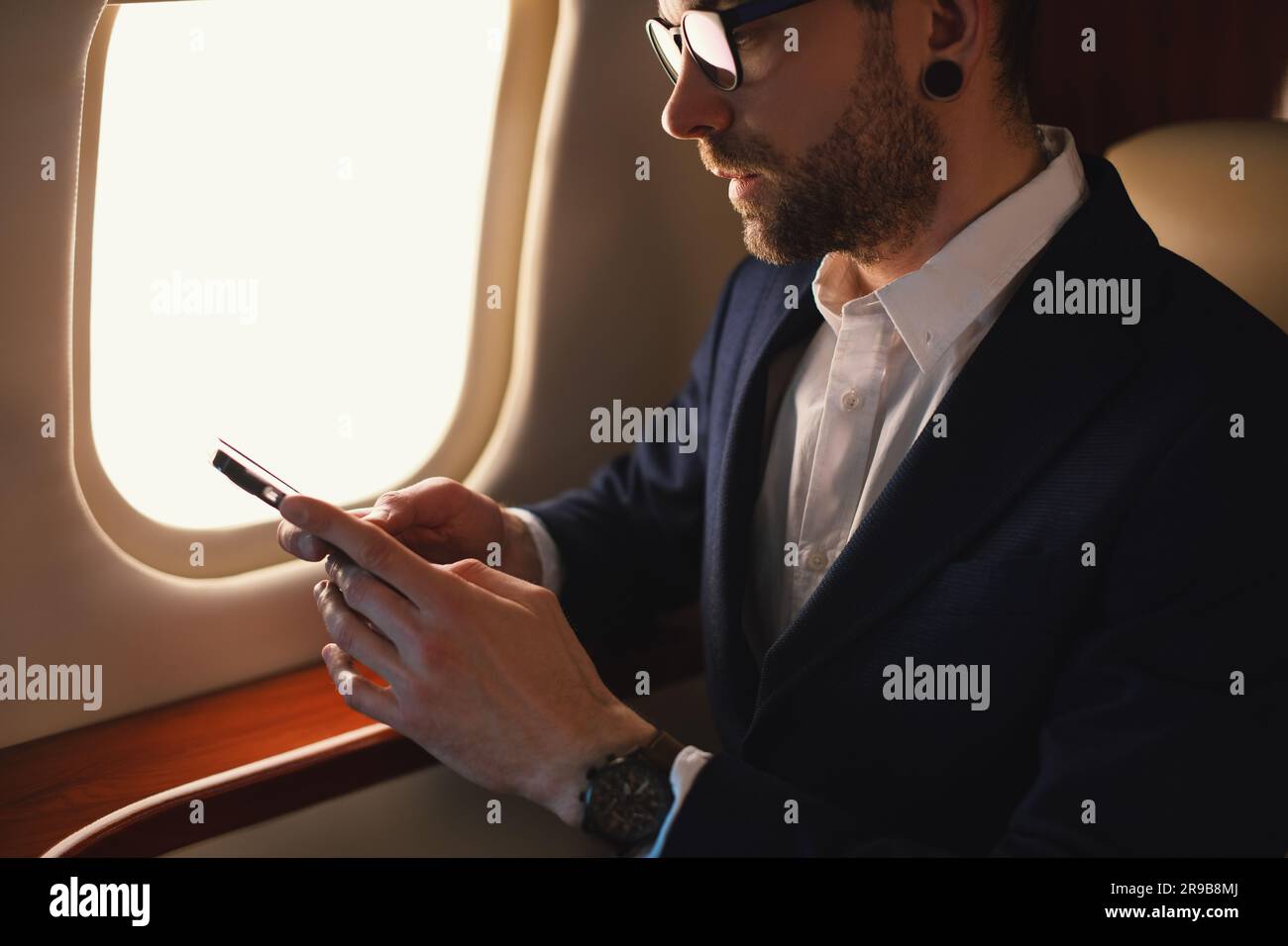 Eleganter CEO junger, selbstbewusster Geschäftsmann mit Brille Mann sitzt in einem erstklassigen Privatjet und arbeitet mit einem modernen Smartphone aus nächster Nähe Stockfoto