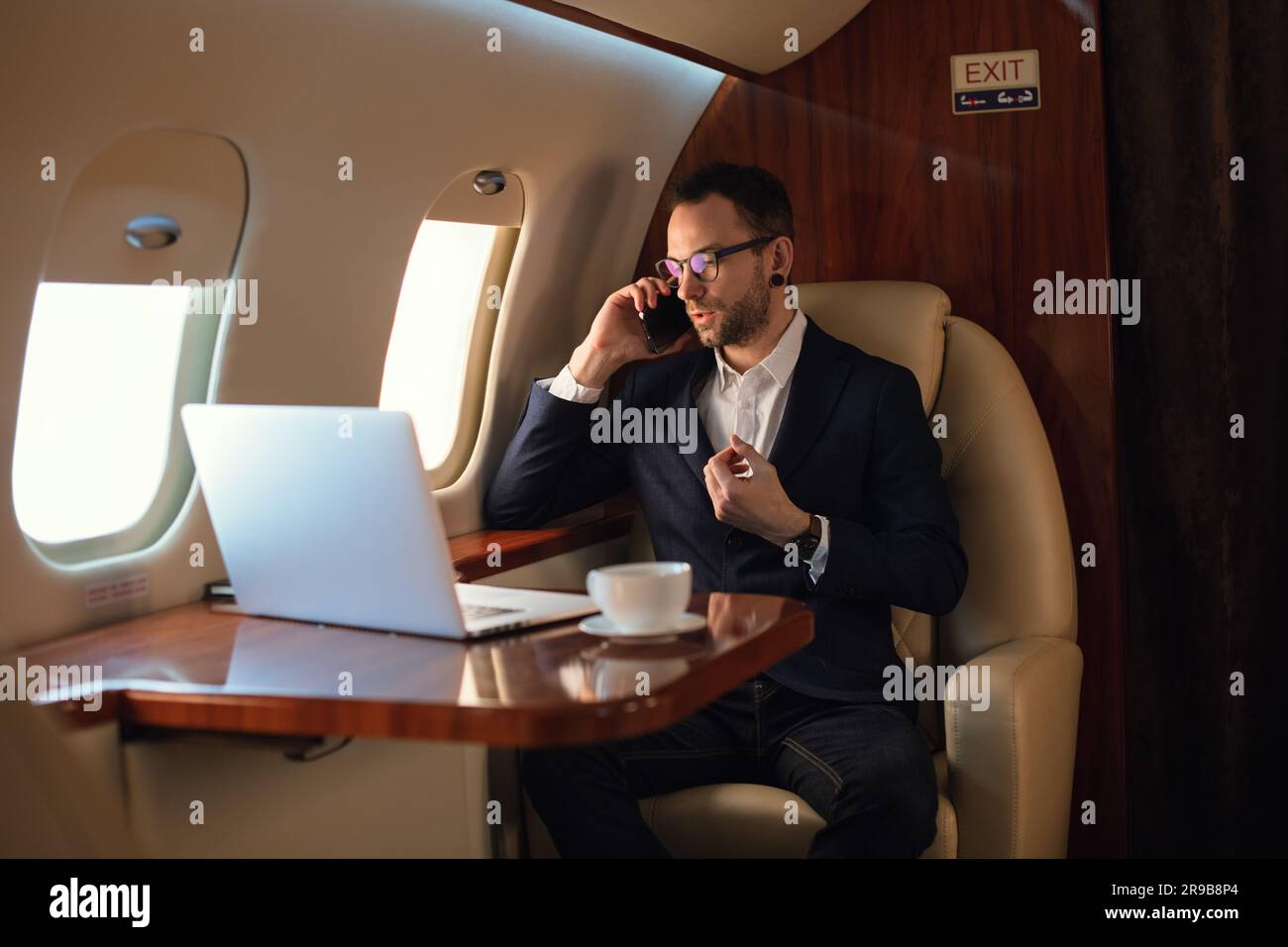 Eleganter junger Geschäftsmann mit Brille, der vor dem Start auf einem Smartphone spricht, in einem Privatflugzeug, Travel Luxury Airline Concept Stockfoto
