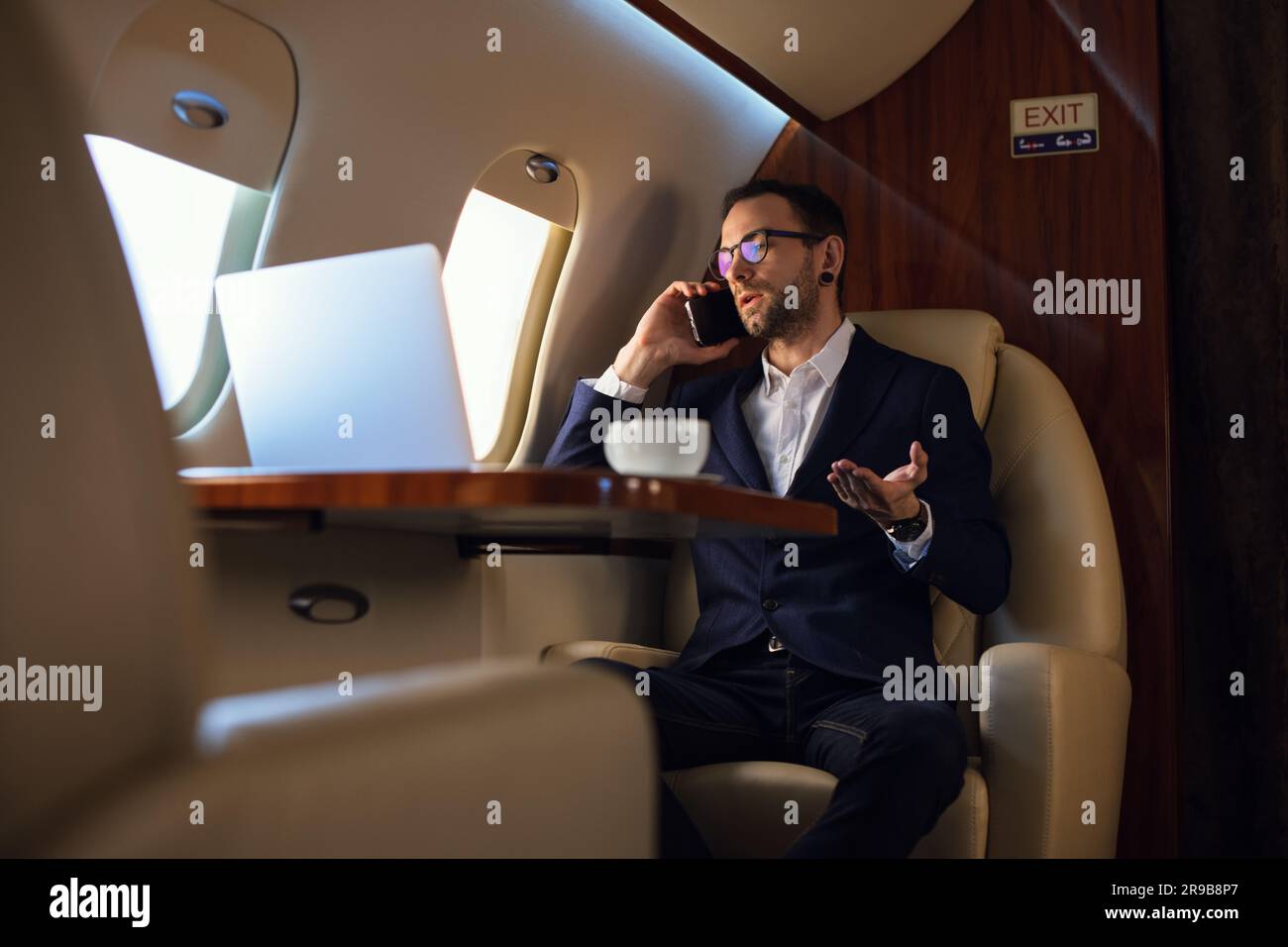 Eleganter junger Geschäftsmann mit Brille, der vor dem Start auf einem Smartphone spricht, in einem Privatflugzeug, Travel Luxury Airline Concept Stockfoto