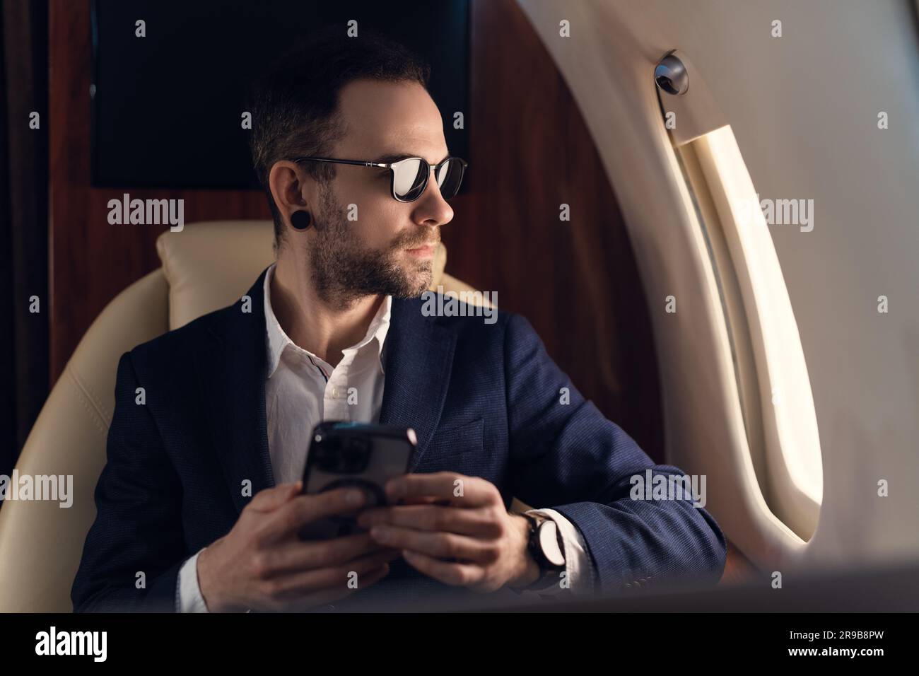 Eleganter junger, selbstbewusster Geschäftsmann mit Sonnenbrille in einem Jackett, der in einem erstklassigen Privatjet sitzt, während er ein modernes Smartphone verwendet Stockfoto