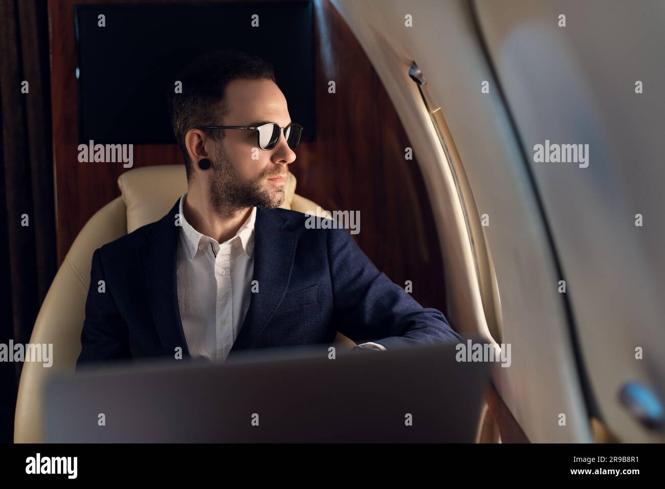 Eleganter, selbstbewusster CEO männlicher Geschäftsmann mit Sonnenbrille in einer blauen Jacke, der in einem erstklassigen Privatflugzeug sitzt und durch ein Fenster schaut Stockfoto