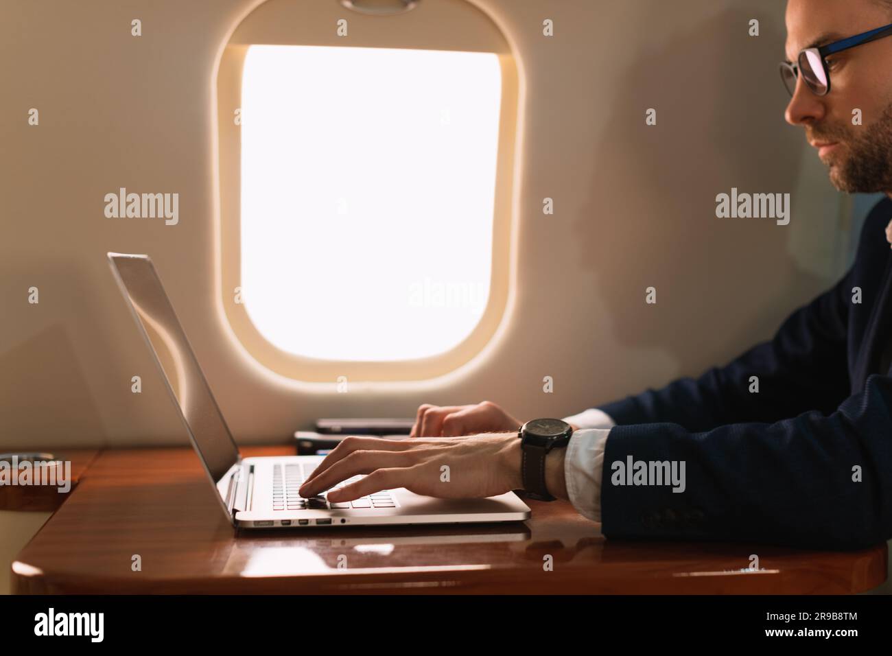 Junger, eleganter Geschäftsführer mit Brille in blauer Jacke und einem modernen Laptop in einem erstklassigen Privatflugzeug mit Jet Side View Stockfoto