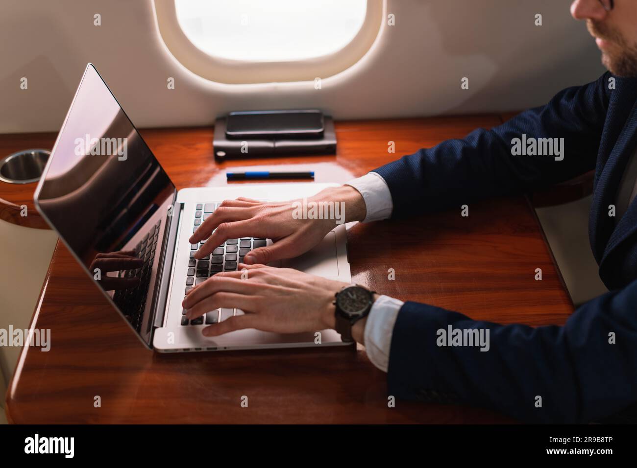 Junger, eleganter Geschäftsführer mit Brille in blauer Jacke und einem modernen Laptop in einem erstklassigen Privatflugzeug mit Jet Side View Stockfoto