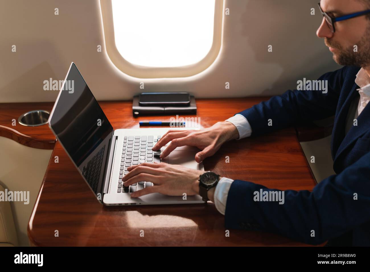 Junger, eleganter Geschäftsführer mit Brille in blauer Jacke und einem modernen Laptop in einem erstklassigen Privatflugzeug mit Jet Side View Stockfoto