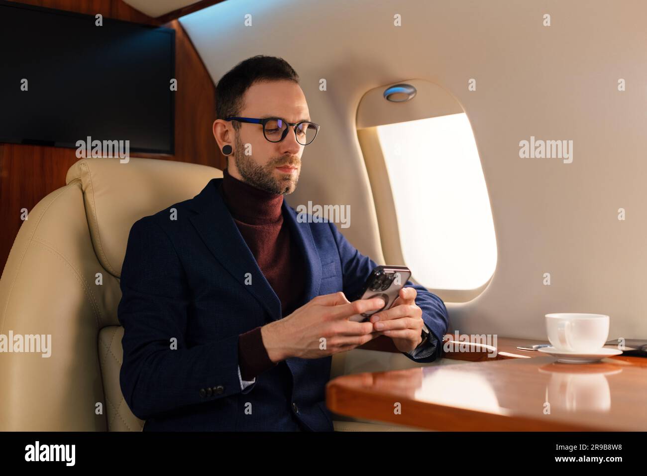Eleganter junger CEO junger Geschäftsmann mit Brille Mann sitzt in einem erstklassigen Privatjet, während er mit einem modernen Smartphone arbeitet Stockfoto
