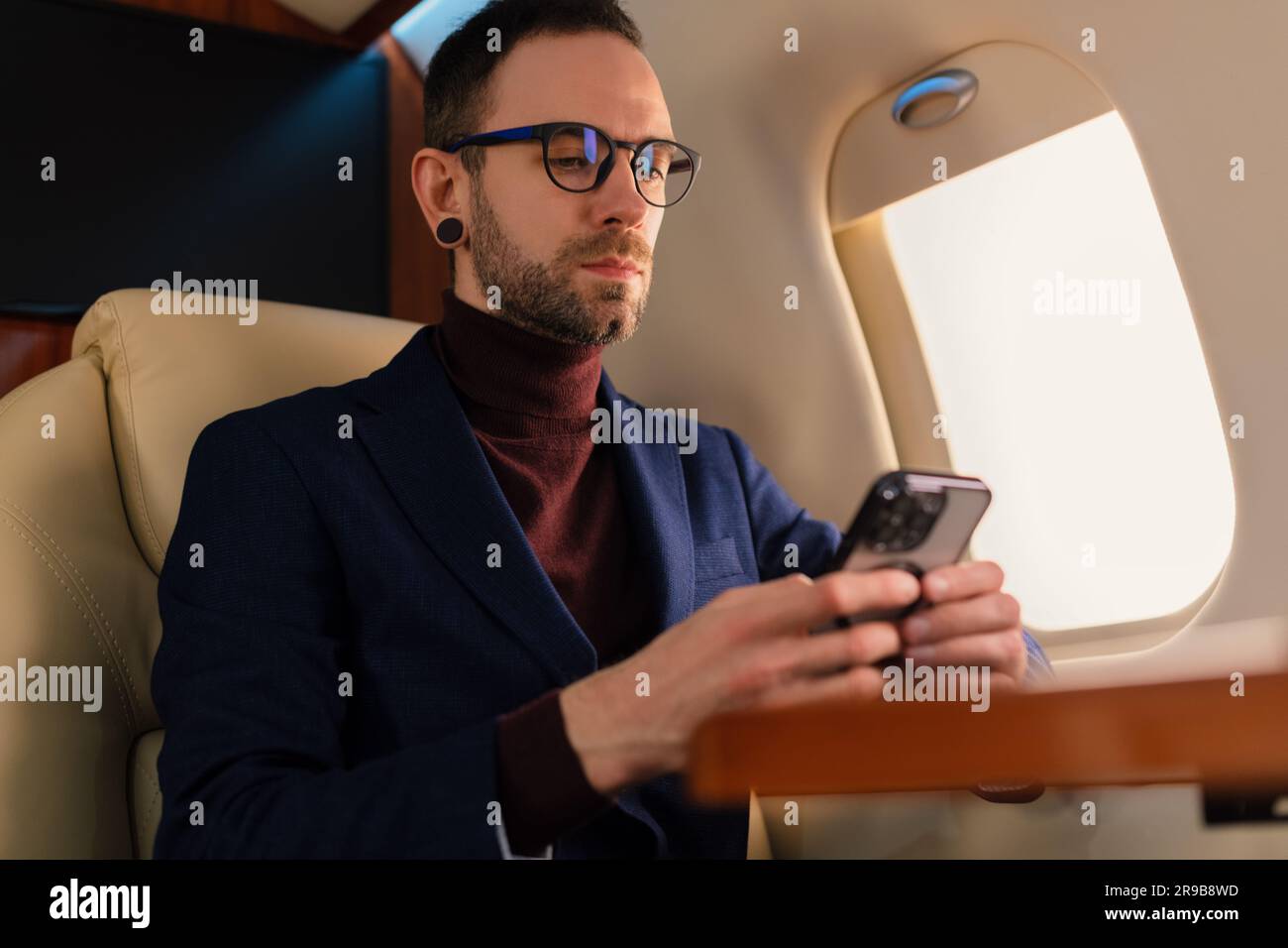 Eleganter junger CEO junger Geschäftsmann mit Brille Mann sitzt in einem erstklassigen Privatjet, während er mit einem modernen Smartphone arbeitet Stockfoto