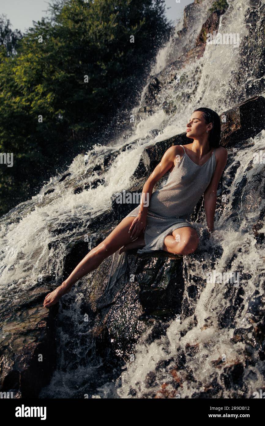 Junge Frau in nassem Kleid, die auf einem Felsen sitzt und mit geschlossenen Augen in der Nähe des Wasserfalls zwischen Wasserströmen, Sprühnebel und Schaum vor dem Hintergrund von Bäumen genießt. Stockfoto
