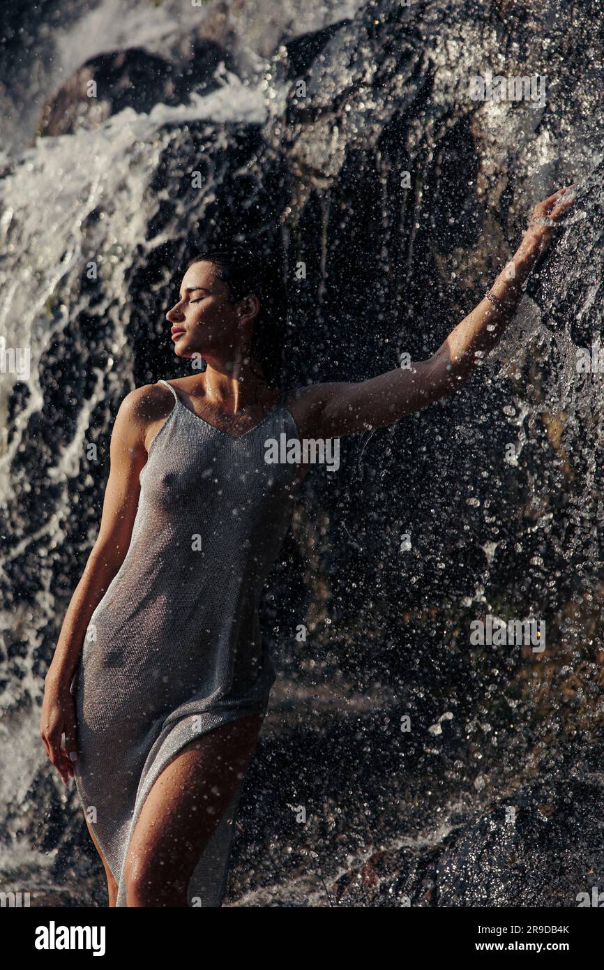 Junge Frau in nassem Kleid steht in der Nähe des Wasserfalls zwischen Wasserströmen, Spray und Schaum. Nahaufnahme. Stockfoto