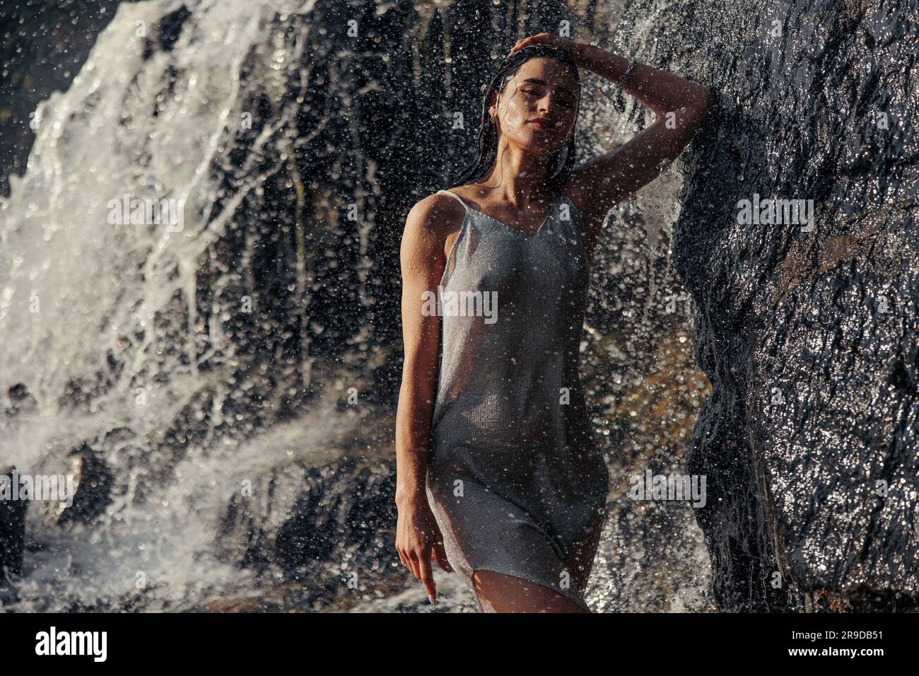 Junge Frau in nassem Kleid steht in der Nähe des Wasserfalls zwischen Wasserströmen, Spray und Schaum. Stockfoto