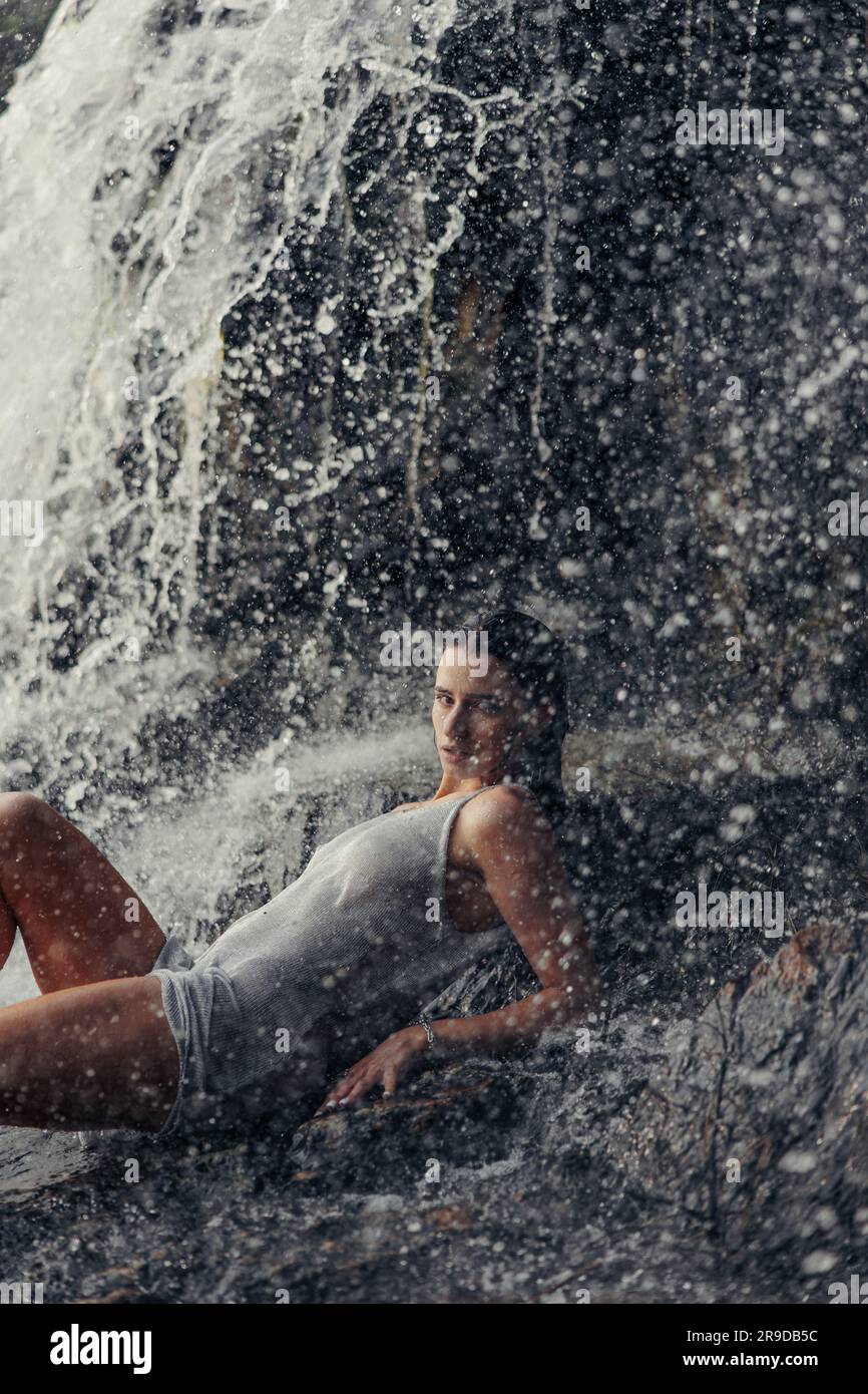 Junge Frau in nassem Kleid, die auf einem Felsen liegt und sich in der Nähe des Wasserfalls zwischen Wasserströmen, Sprühnebel und Schaum befindet. Stockfoto