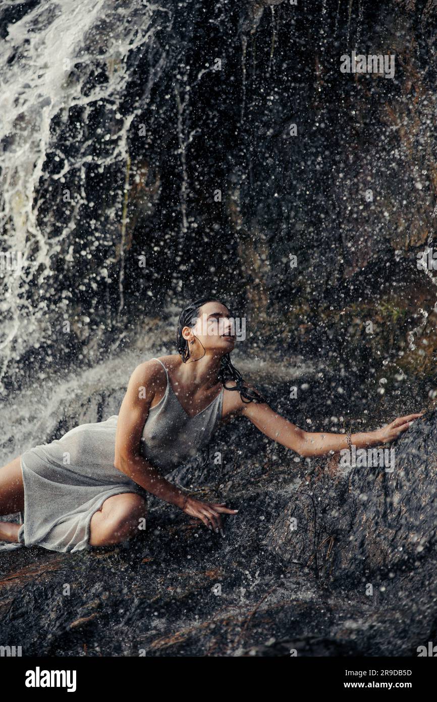 Junge Frau in nassem Kleid, die auf einem Felsen liegt und mit geschlossenen Augen in der Nähe des Wasserfalls zwischen Wasserströmen, Sprühnebel und Schaum genießt. Stockfoto
