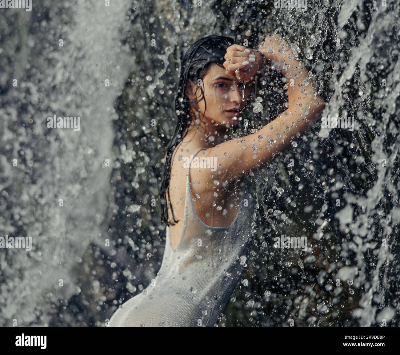 Porträt einer jungen Frau in nassem Kleid, die im Wasserfall zwischen Wasserströmen, Sprühnebel und Tropfen steht. Nahaufnahme. Stockfoto