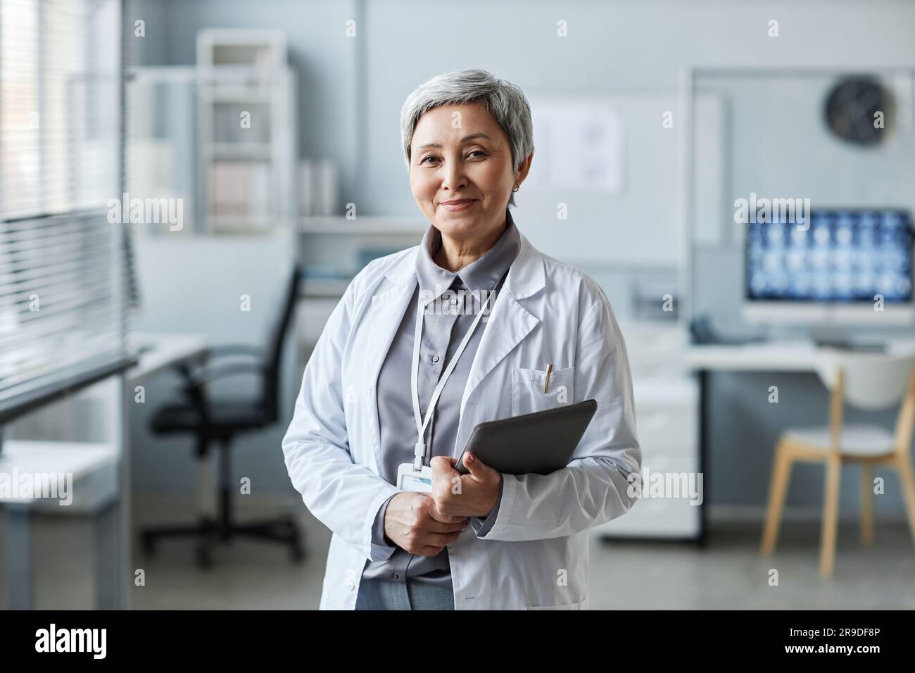 Glückliche reife Ärztin mit Tablet steht vor der Kamera in der Arztpraxis und schaut Sie gegen die Arbeitsplätze von Kollegen Stockfoto