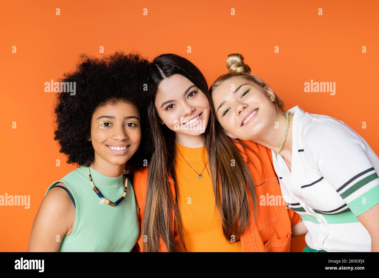 Porträt von lächelnden und multiethnischen Teenagern mit hellem Make-up, die in die Kamera schauen, während sie zusammen posieren, isoliert auf orangefarbenen, trendigen Outfits und Stockfoto