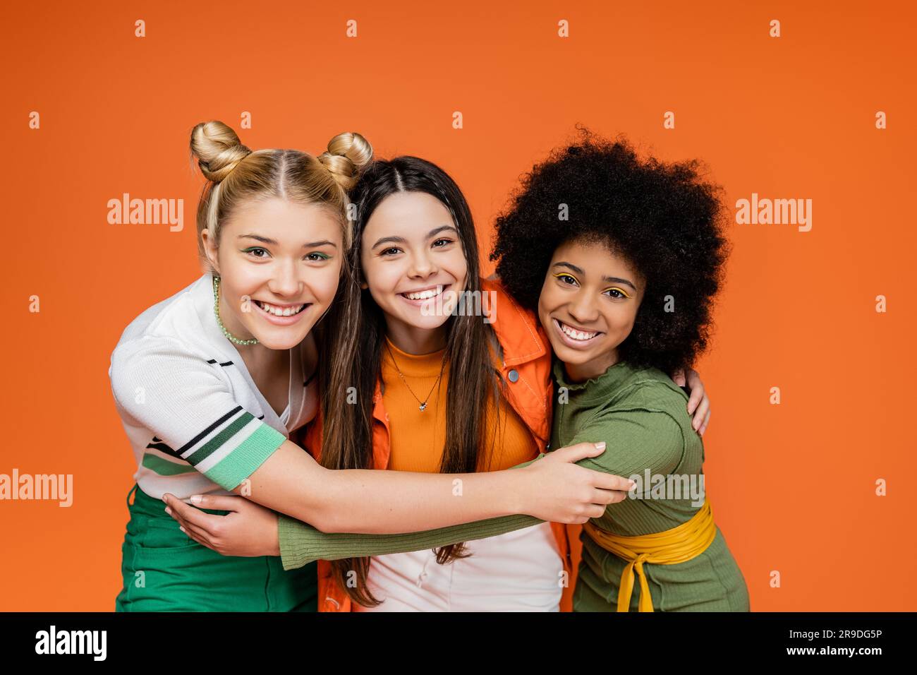 Porträt von positiven und interrassistischen Teenager-Freundinnen in trendigen Outfits, die sich umarmen und in die Kamera schauen, während sie isoliert auf Orange stehen, Stockfoto