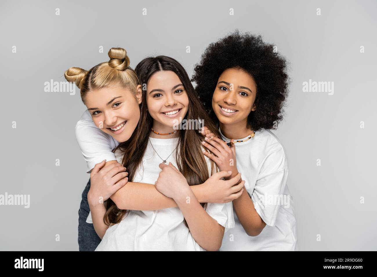 Porträt positiver und interrassistischer Teenager-Mädchen in weißen T-Shirts, die brünette Freundin umarmen und auf die Kamera schauen, isoliert auf graue, Jugendliche m Stockfoto