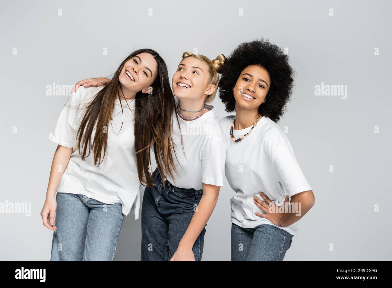 Multiethnische Teenager-Freundinnen in Jeans und legeren weißen T-Shirts posieren, während sie sich umarmen und zusammenstehen, isoliert auf grauen Jugendmodellen und Stockfoto