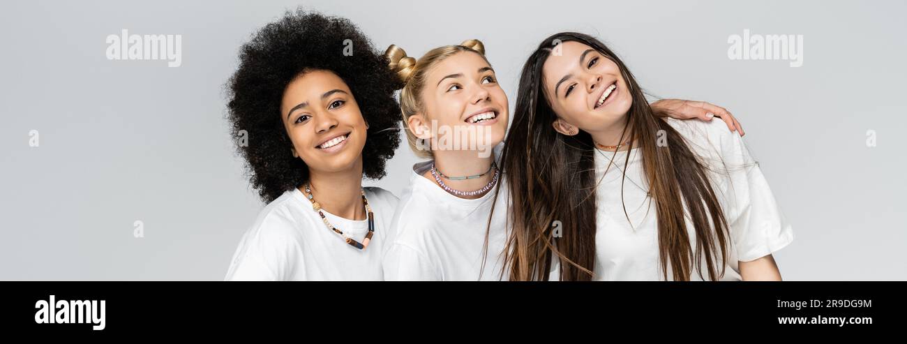Positive und multiethnische Teenager-Freundinnen in legeren weißen T-Shirts, die sich umarmen und in die Kamera schauen, während sie isoliert auf einem grauen Jugendmodell stehen Stockfoto